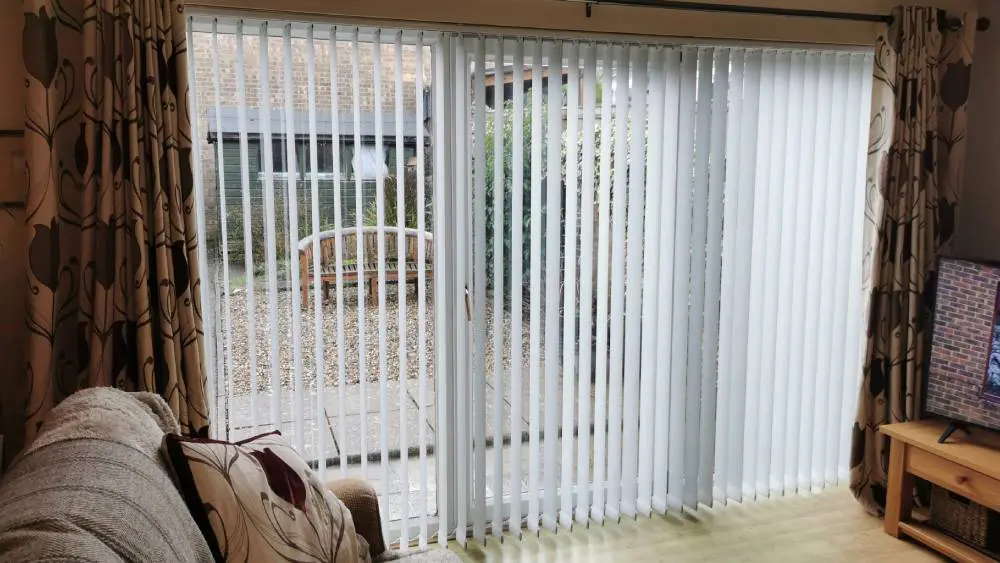 White vertical blinds fitted across bi-fold patio doors in a living room, partially open to show the garden
