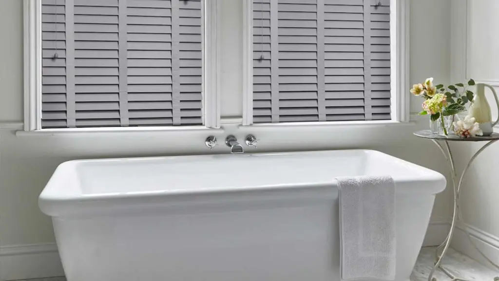 Grey painted Venetian blinds in an elegant bathroom with a freestanding bath and fresh flowers